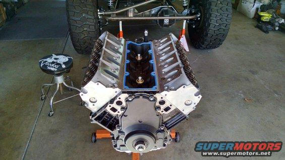 V8 engine on an orange stand, with a Jeep's rear tire and suspension visible behind it. A stool with 'Voodoo' text is to the left