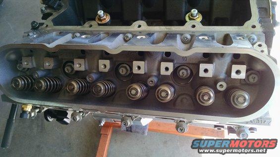 Close-up of an engine cylinder head with valves and springs visible. Some bolts and numbers are present on the metal