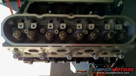 Engine cylinder head with visible valves and springs, mounted on an engine stand. Text 'SUPERMOTORS' visible