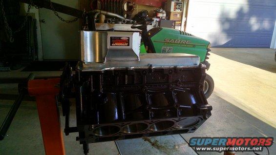 Black engine block on an orange engine stand, with a silver oil pan and intake manifold attached. A green lawnmower is in the background
