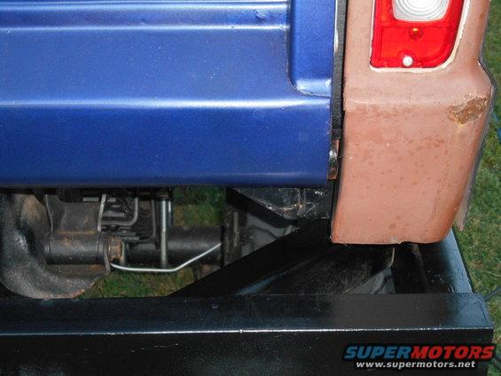 Close-up of a blue rear quarter panel and brown fender with a rusty spot. Suspension components and part of a black frame are visible below