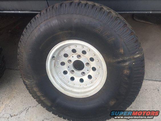 Close-up of a large off-road tire mounted on a silver multi-hole wheel. Some text is visible on the tire sidewall