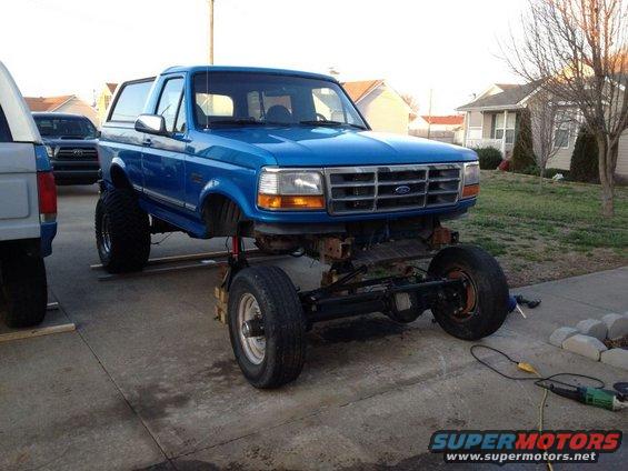 blue Ford Bronco with front end raised on blocks, showing suspension work. Another vehicle is parked to the left