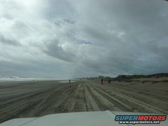 Driving on a sandy beach with tire tracks and cloudy skies. Some people are visible in the distance