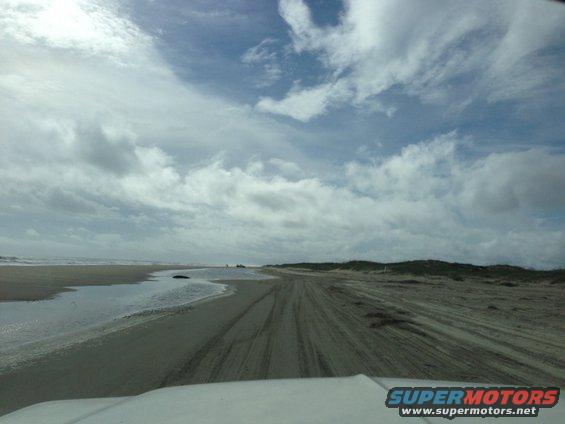 View from inside a vehicle driving on a sandy beach with tire tracks and ocean in the distance