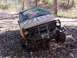 1983 Ford Bronco - This damaged off-road vehicle is resting on uneven terrain in a wooded area, with its front end visible.
