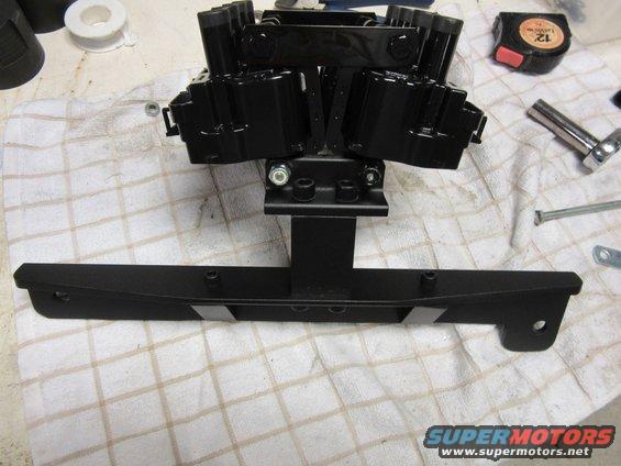 Black engine ignition coil pack assembly mounted on a bracket, with tools and tape measure nearby