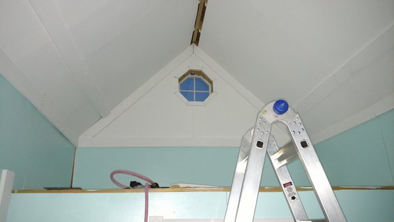Interior of a room with light blue walls, white sloped ceiling, an octagonal window, and a silver extension ladder