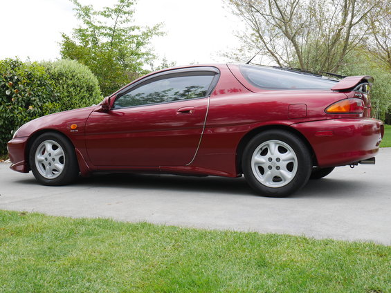 Red 1993 Mazda MX-3 coupe, side view, parked on concrete driveway with grass and trees in background