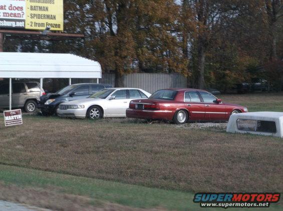 Red 1995 Mercury Grand Marquis parked next to a silver sedan and a dark SUV under a carport