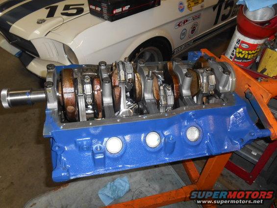 Engine block on stand, with crankshaft installed. White race car with blue stripes and number 15 in background