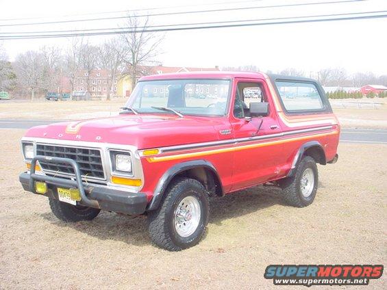 Red 1978 Ford Bronco with chrome grille guard and yellow fog lights, parked on dry grass