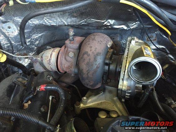 Turbocharger assembly with rusty exhaust housing and metal intake pipe, surrounded by engine wiring and insulation