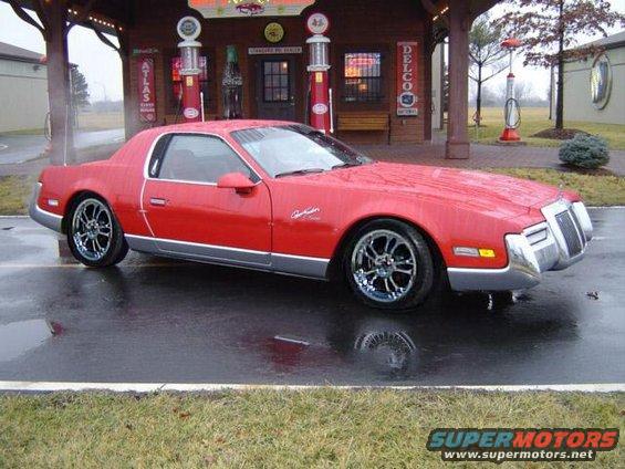 Red coupe with chrome wheels parked on a wet asphalt lot in front of a vintage gas station