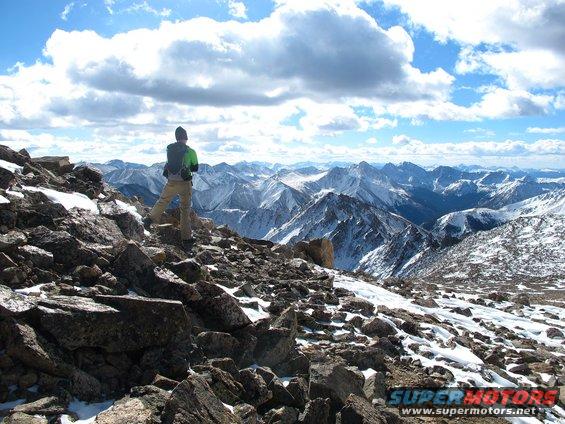Hiker on rocky mountain peak overlooking snow-covered ranges under a cloudy sky