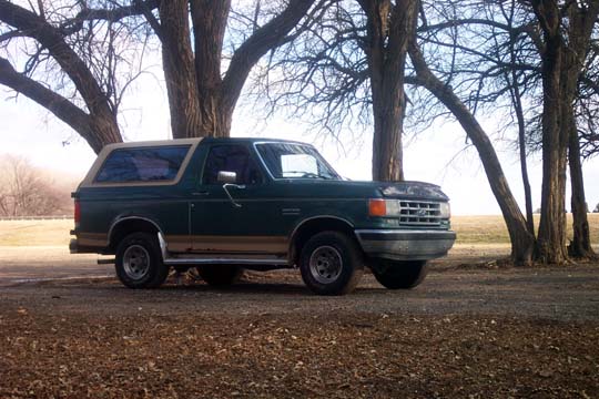 Green 1987 Ford Bronco parked under trees, tan trim, aftermarket wheels