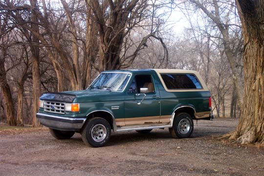 Green Ford Bronco with tan trim parked on a dirt road, trees in background