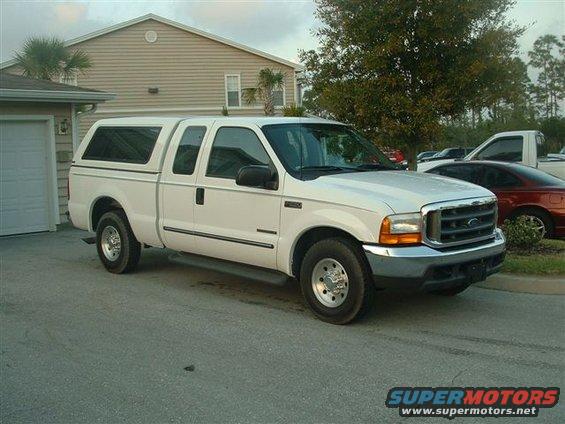 White pickup truck with a camper shell parked in front of a house. Another red car is parked behind it