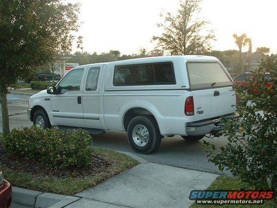 White pickup truck with a camper shell, seen from the side at a slight rear angle. The truck has four doors and appears to be in good condition