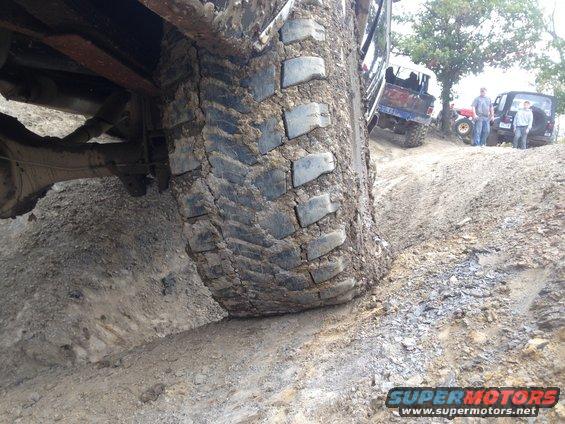 Off-road vehicles on a dirt hill, close-up of a muddy tire. Other vehicles and people visible in the background