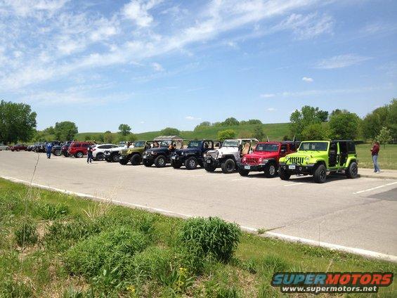Line of Jeeps parked in a lot on a sunny day. Some people are standing around the vehicles