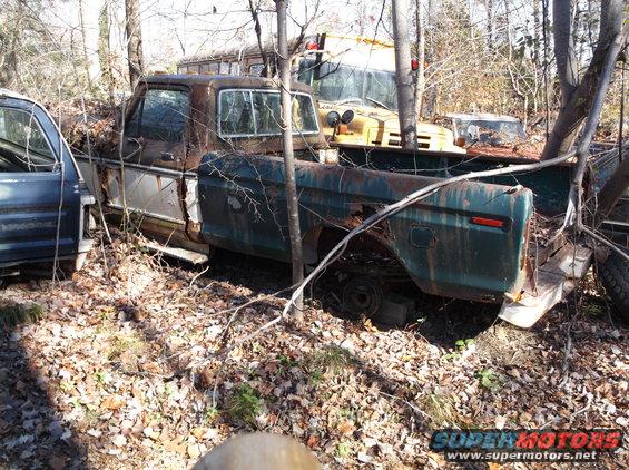 Rusty green and brown pickup truck in a wooded junkyard, side view