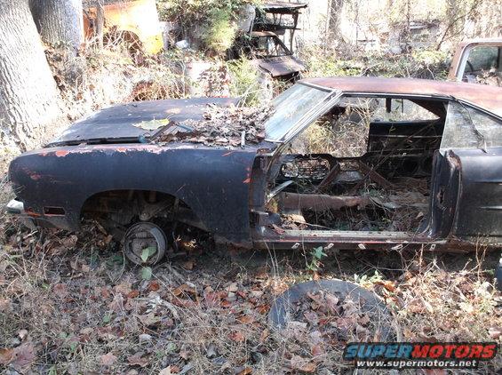 Old muscle car, looks like a 1970 model, sitting in a junkyard with leaves and debris covering it. Hood is off, doors open, interior is gutted