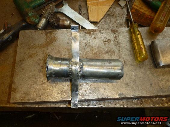 Welded metal component with a flared end and a cross-shaped bracket, resting on a metal plate on a workbench with tools
