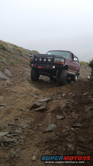 Maroon Ford Bronco with aftermarket bumper and lights driving on a rocky, dirt trail