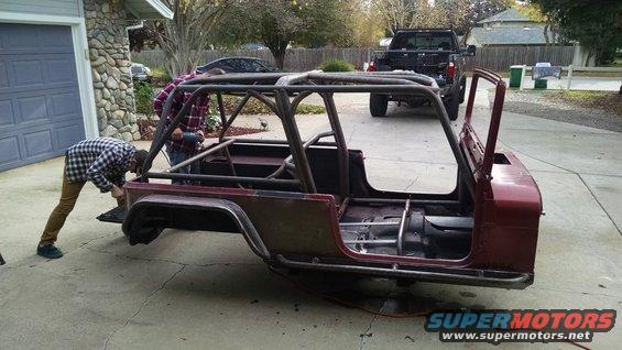 Two guys working on a red Jeep CJ body with a custom roll cage installed. A black pickup truck is parked behind it