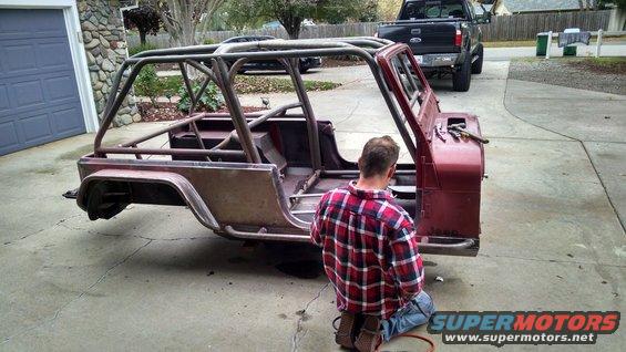 Red Jeep CJ body with custom roll cage, missing doors and windshield, a person kneels beside it