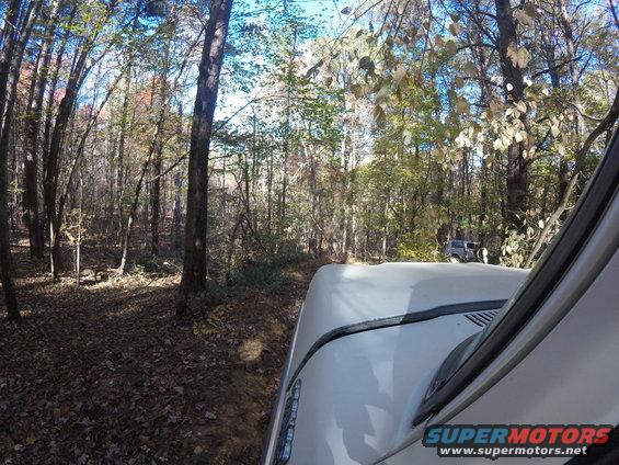 White SUV driving on a forest road, another SUV visible in the distance