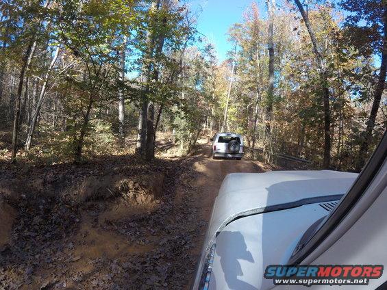 Two SUVs on a dirt road in a forest, one behind the other. Watermark 'SUPERMOTORS.NET' at bottom right