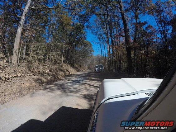 White 1979 Ford Bronco driving on a dirt road, with another vehicle ahead