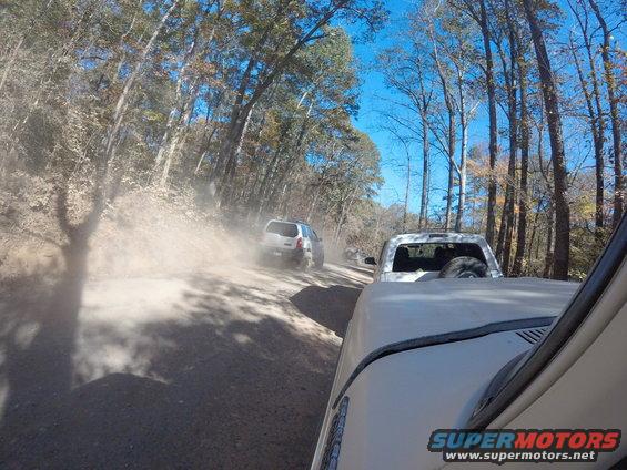 Two off-road vehicles driving on a dusty dirt road through trees. One vehicle is in the foreground, partially visible