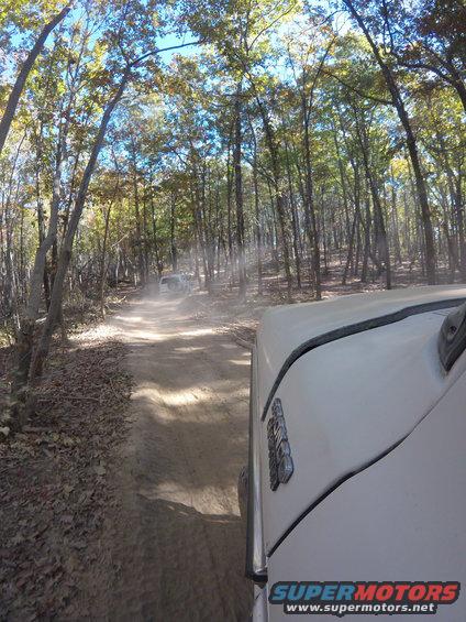 White SUV driving on a dusty forest road, another white SUV in the distance