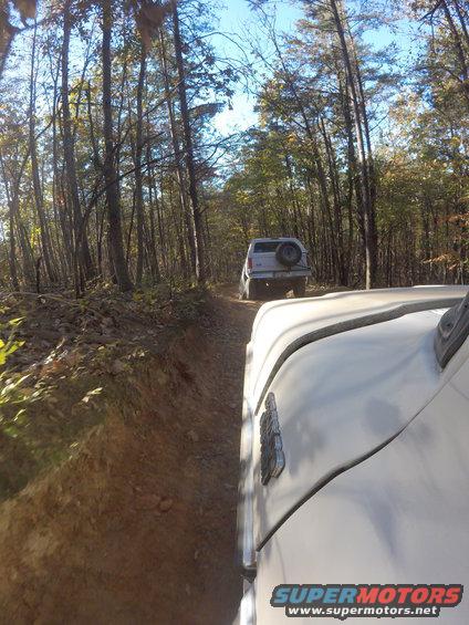 Two 1979 Ford Broncos driving on a wooded dirt road