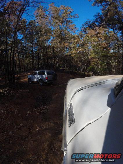Two SUVs driving on a dirt road through a forest. One SUV is in the foreground, partially visible, and another is in the background