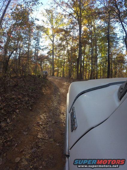 1979 Ford Bronco driving on a dirt road in a forest with another SUV ahead