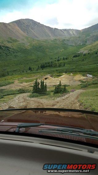 Red dashboard and windshield wipers in foreground, overlooking a vast mountain valley with a dirt road