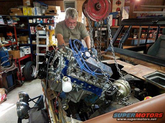 Man lowering a blue engine into a vehicle bay with a hoist