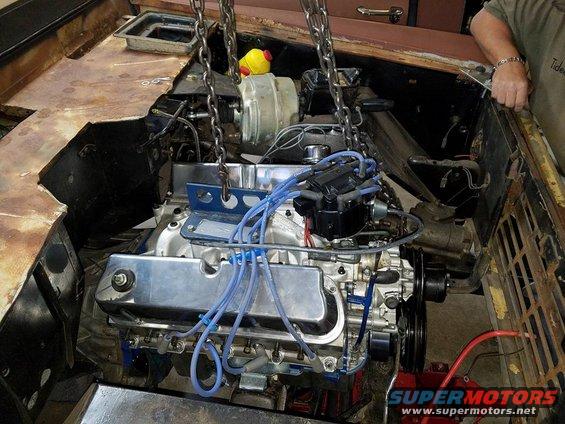 Engine being lowered into a vehicle chassis with chains. A person is visible on the right