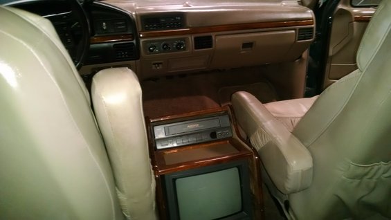 Tan leather captain's chairs face a wood-trimmed console with a VCR and CRT television. Dashboard and steering wheel visible