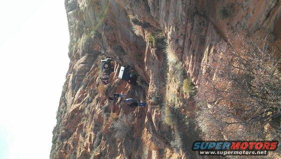 Multiple off-road vehicles precariously positioned on a steep, rocky cliff face under a bright sky
