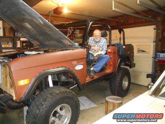 Man reading a book in a 1972 Ford Bronco with the hood up in a garage