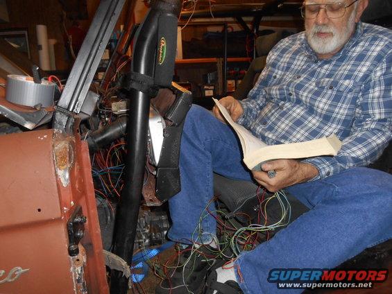 Man in blue jeans and plaid shirt reads manual amidst tangled wires inside a rusty vehicle