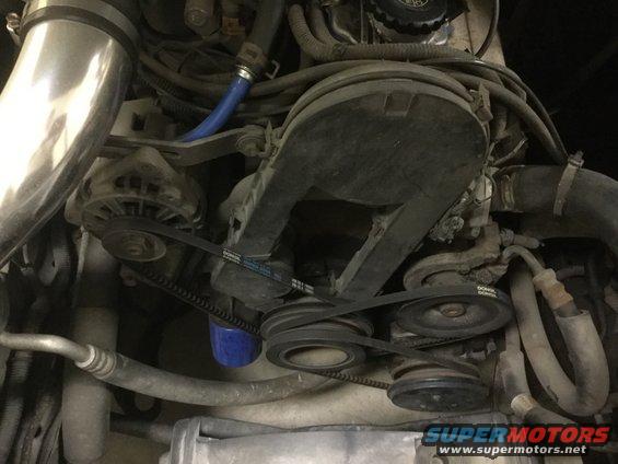 Engine bay with alternator, belts, pulleys, and blue oil filter. Some hoses and wiring are visible