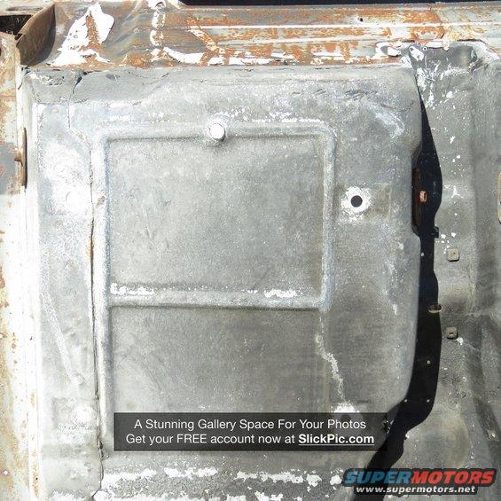 Undercarriage of a damaged vehicle, showing a metal panel with a raised rectangular section and a circular hole
