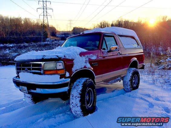 Red Ford Bronco covered in snow, parked in a snowy field with power lines and trees in the background at sunset