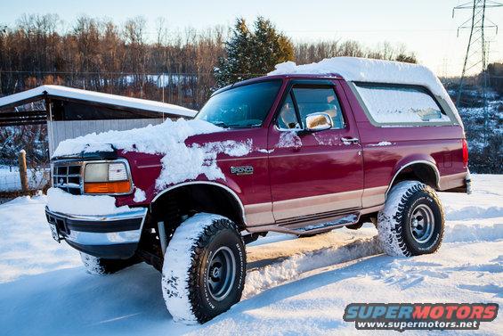 Maroon Ford Bronco with a tan stripe, covered in snow, parked in a snowy outdoor area